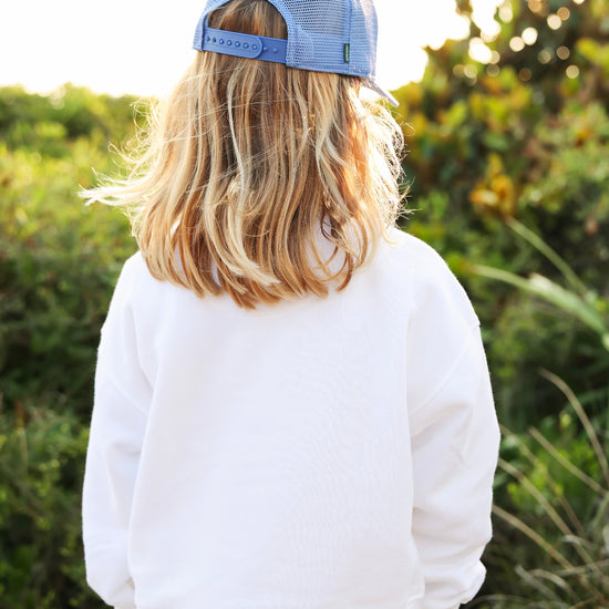 A child viewed from behind wearing a light blue trucker cap and a white sweatshirt, standing outdoors in front of a blurred green background.