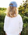 A child viewed from behind wearing a light blue trucker cap and a white sweatshirt, standing outdoors in front of a blurred green background.