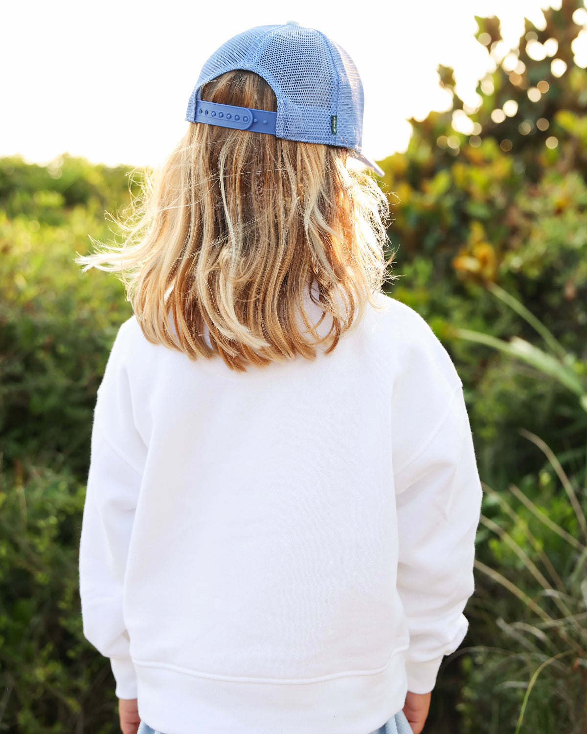 A child viewed from behind wearing a light blue trucker cap and a white sweatshirt, standing outdoors in front of a blurred green background.