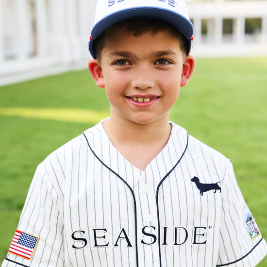 A young boy smiling while wearing a white pinstripe baseball jersey and matching baseball cap with 'SEASIDE' printed on it. The jersey features a black dachshund logo on the left chest and a small American flag patch on the right sleeve, set against a blurred green grass background.