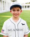 A young boy smiling while wearing a white pinstripe baseball jersey and matching baseball cap with 'SEASIDE' printed on it. The jersey features a black dachshund logo on the left chest and a small American flag patch on the right sleeve, set against a blurred green grass background.