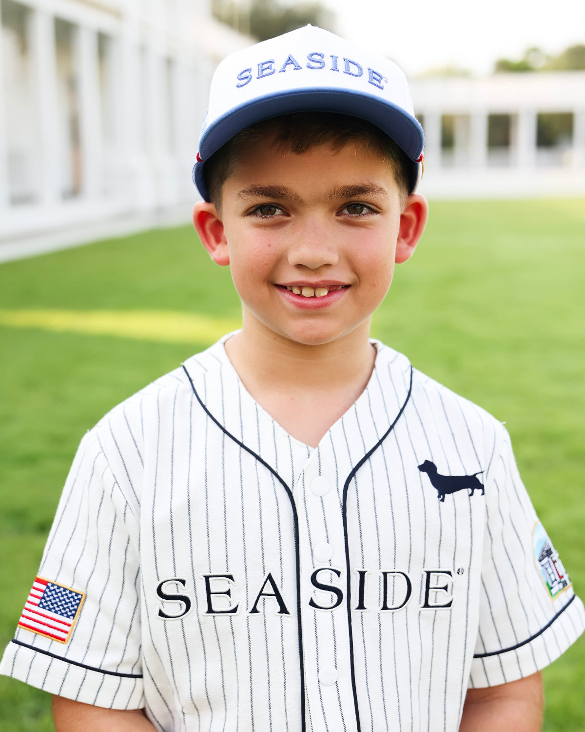 A young boy smiling while wearing a white pinstripe baseball jersey and matching baseball cap with 'SEASIDE' printed on it. The jersey features a black dachshund logo on the left chest and a small American flag patch on the right sleeve, set against a blurred green grass background.