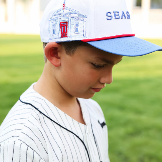 A young boy wearing a white and blue baseball cap with a red trim and a seaside post office patch, paired with a white pinstripe baseball jersey, standing on a green grass field.