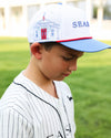 A young boy wearing a white and blue baseball cap with a red trim and a seaside post office patch, paired with a white pinstripe baseball jersey, standing on a green grass field.