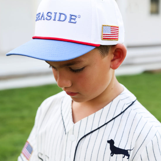 A young boy wearing a white baseball cap with blue brim and 'SEASIDE' embroidered on the front, paired with a white pinstripe baseball jersey. The photo is taken outdoors on a grassy lawn. 