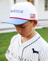 A young boy wearing a white baseball cap with blue brim and 'SEASIDE' embroidered on the front, paired with a white pinstripe baseball jersey. The photo is taken outdoors on a grassy lawn. 