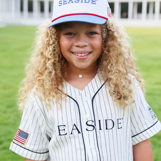 A young girl with long curly blonde hair smiles while wearing a white pinstripe baseball jersey and a matching white 'SEASIDE' cap on a green grass field.
