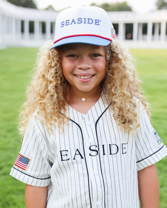 A young girl with long curly blonde hair smiles while wearing a white pinstripe baseball jersey and a matching white 'SEASIDE' cap on a green grass field.