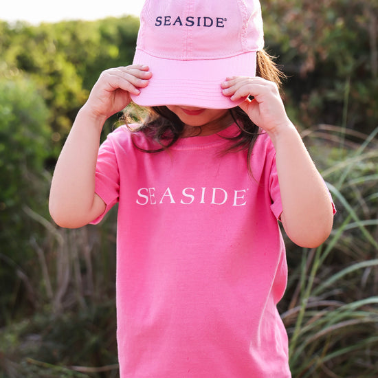 A child wearing a pink baseball cap with 'SEASIDE' embroidered on the front and a matching pink t-shirt with 'SEASIDE' printed across the chest, holding the brim of the hat in front of their face against an outdoor grassy background.