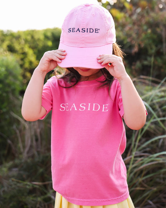 A child wearing a pink baseball cap with 'SEASIDE' embroidered on the front and a matching pink t-shirt with 'SEASIDE' printed across the chest, holding the brim of the hat in front of their face against an outdoor grassy background.