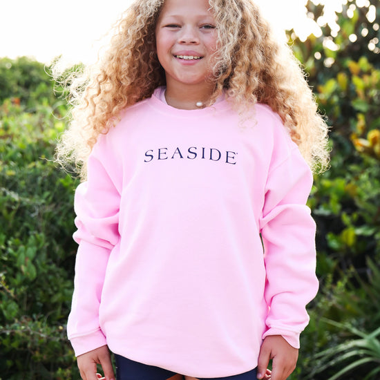 A young girl with long, curly blonde hair stands outdoors wearing a light pink crewneck sweatshirt with 'SEASIDE' printed in navy blue across the chest, paired with dark blue shorts.