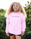 A young girl with long, curly blonde hair stands outdoors wearing a light pink crewneck sweatshirt with 'SEASIDE' printed in navy blue across the chest, paired with dark blue shorts.