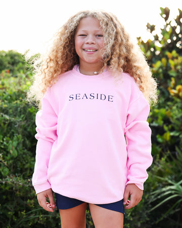 A young girl with long, curly blonde hair stands outdoors wearing a light pink crewneck sweatshirt with 'SEASIDE' printed in navy blue across the chest, paired with dark blue shorts.