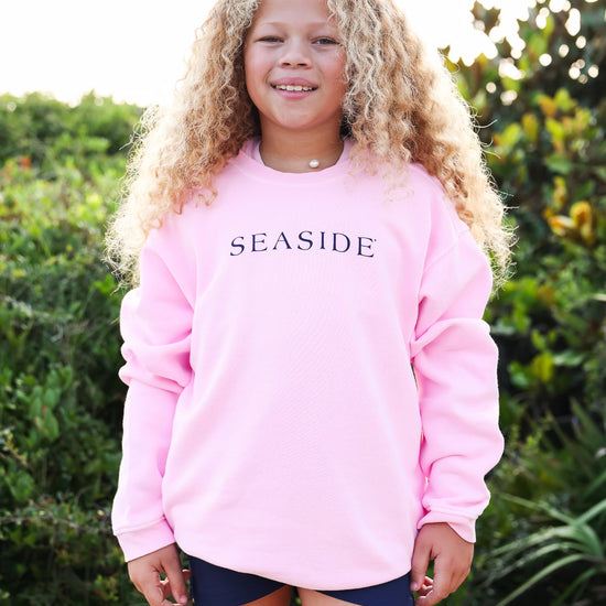 A young girl with long curly blonde hair stands outdoors wearing a light pink crewneck sweatshirt with the word 'SEASIDE' printed in navy blue across the chest, paired with dark navy shorts.