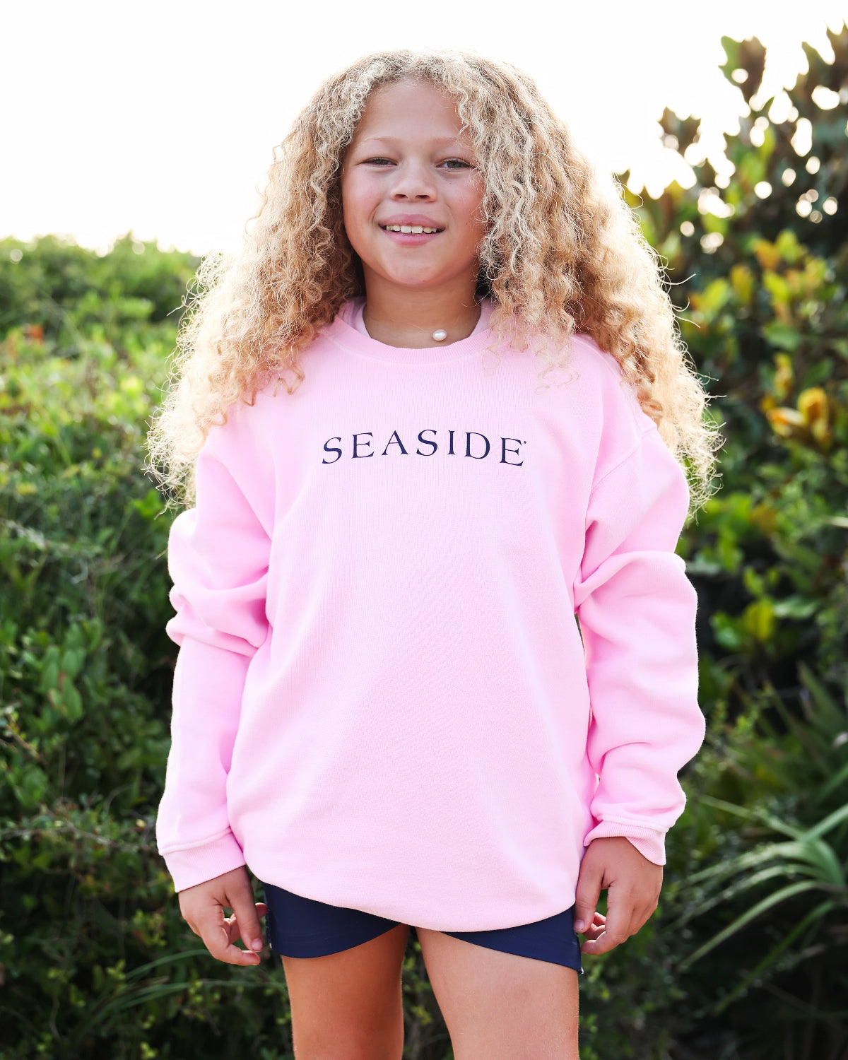 A young girl with long curly blonde hair stands outdoors wearing a light pink crewneck sweatshirt with the word 'SEASIDE' printed in navy blue across the chest, paired with dark navy shorts.