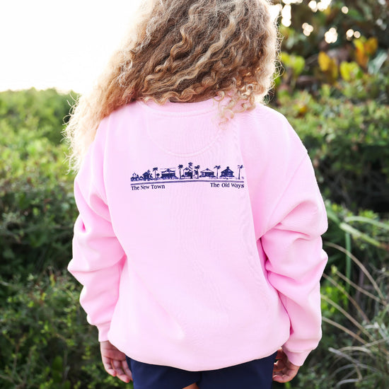 A child viewed from behind wearing a light pink crewneck sweatshirt with a dark blue graphic print across the upper back, paired with navy shorts, set against a blurred outdoor background of green foliage.
