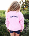 A child viewed from behind wearing a light pink crewneck sweatshirt with a dark blue graphic print across the upper back, paired with navy shorts, set against a blurred outdoor background of green foliage.