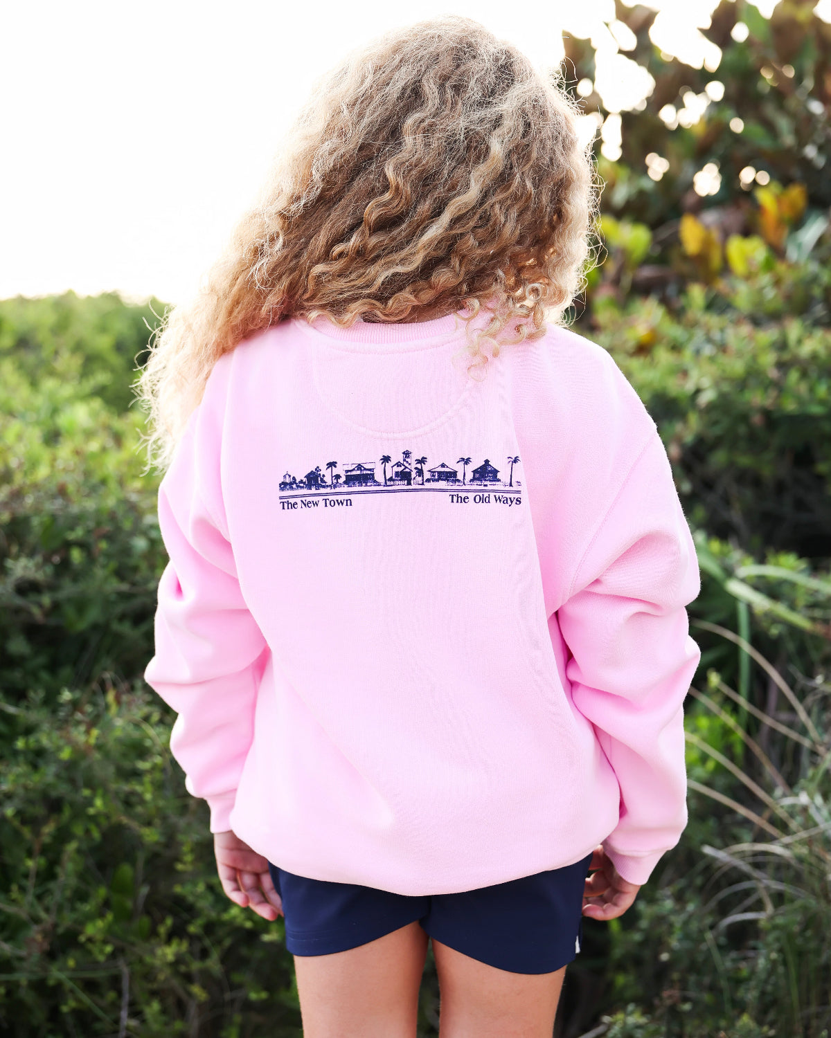 A child viewed from behind wearing a light pink crewneck sweatshirt with a dark blue graphic print across the upper back, paired with navy shorts, set against a blurred outdoor background of green foliage.
