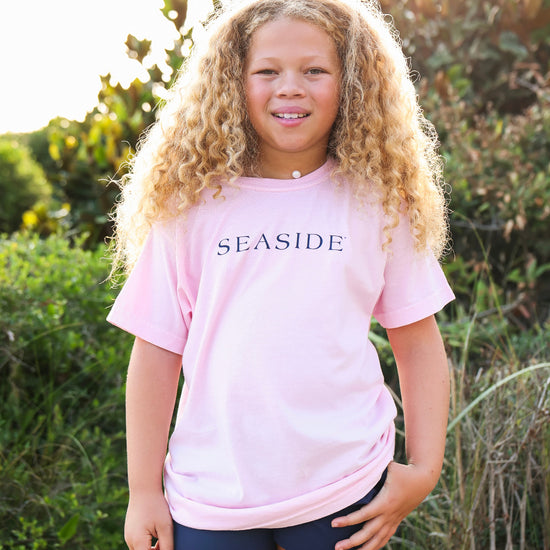 A young girl with long curly blonde hair stands outdoors wearing a light pink t-shirt with 'SEASIDE' printed in navy blue across the chest, paired with dark navy shorts.