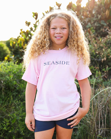 A young girl with long curly blonde hair stands outdoors wearing a light pink t-shirt with 'SEASIDE' printed in navy blue across the chest, paired with dark navy shorts.