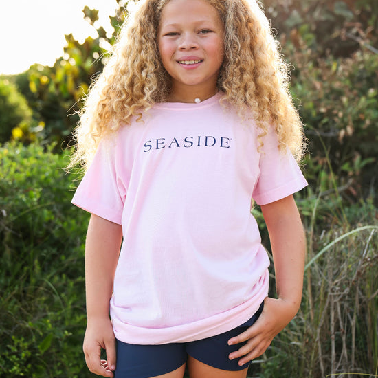 A young girl with long curly blonde hair stands outdoors wearing a light pink short-sleeve t-shirt with 'SEASIDE' printed in navy blue across the chest, paired with dark navy shorts.