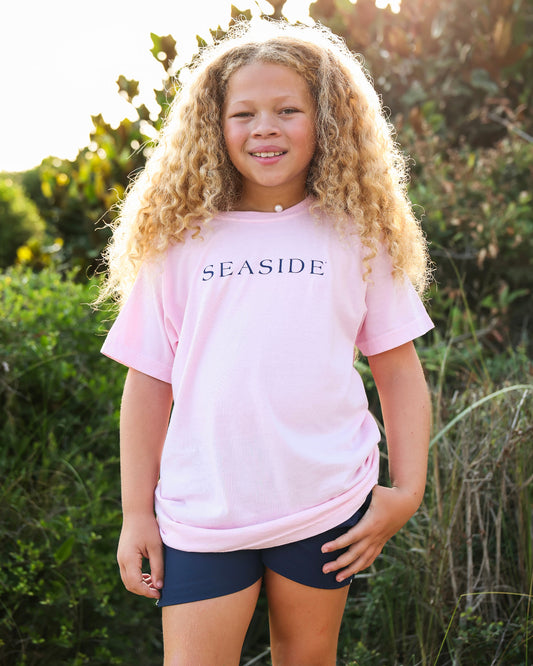 A young girl with long curly blonde hair stands outdoors wearing a light pink short-sleeve t-shirt with 'SEASIDE' printed in navy blue across the chest, paired with dark navy shorts.