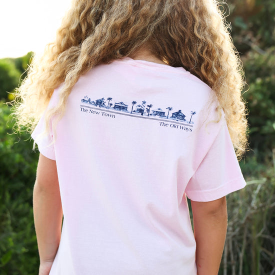 A person with long curly blonde hair seen from behind, wearing a light pink short-sleeve T-shirt with a dark blue graphic and text across the upper back. The background is an outdoor setting with green foliage and warm sunlight.