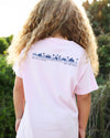 A person with long curly blonde hair seen from behind, wearing a light pink short-sleeve T-shirt with a dark blue graphic and text across the upper back. The background is an outdoor setting with green foliage and warm sunlight.