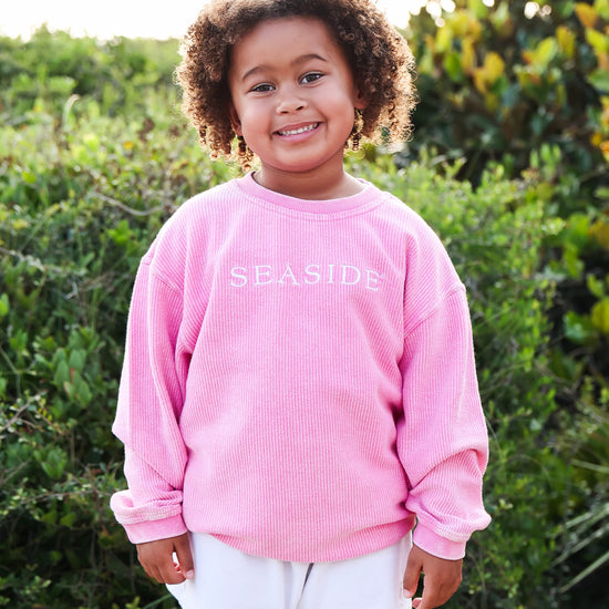 A young girl with curly brown hair stands outdoors in front of green bushes, wearing a pink corded sweatshirt with 'SEASIDE' printed in white across the chest and white shorts.