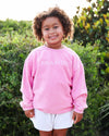 A young girl with curly brown hair stands outdoors in front of green bushes, wearing a pink corded sweatshirt with 'SEASIDE' printed in white across the chest and white shorts.