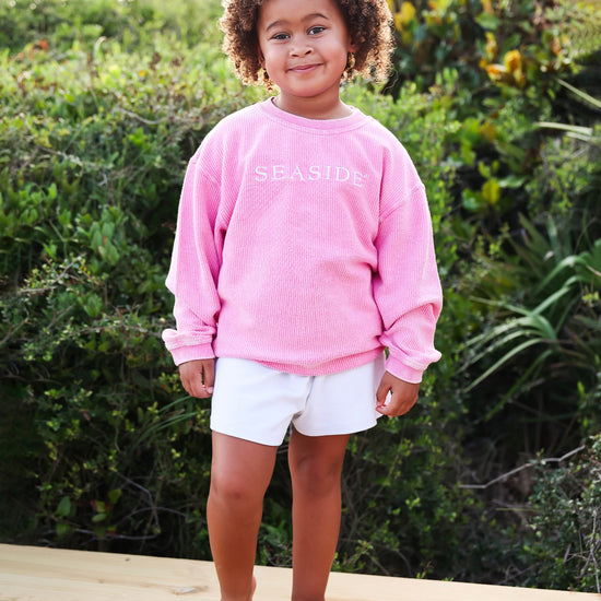 A young child with curly hair stands barefoot on a wooden deck wearing a pink corded crewneck sweatshirt with 'SEASIDE' printed in white across the chest, paired with white shorts, set against a lush green outdoor background.