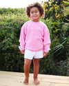 A young child with curly hair stands barefoot on a wooden deck wearing a pink corded crewneck sweatshirt with 'SEASIDE' printed in white across the chest, paired with white shorts, set against a lush green outdoor background.