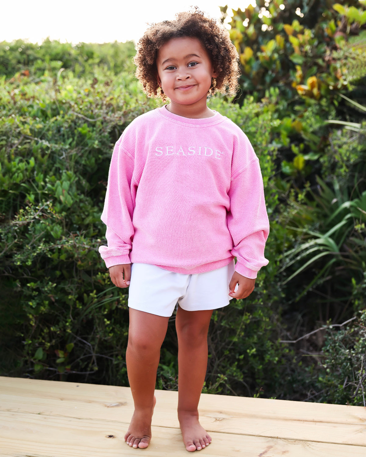 A young child with curly hair stands barefoot on a wooden deck wearing a pink corded crewneck sweatshirt with 'SEASIDE' printed in white across the chest, paired with white shorts, set against a lush green outdoor background.