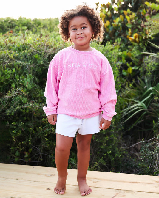A young child with curly hair stands barefoot on a wooden deck wearing a pink corded crewneck sweatshirt with 'SEASIDE' printed in white across the chest, paired with white shorts, set against a lush green outdoor background.