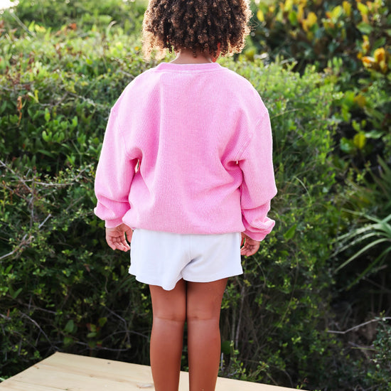 A child standing on a wooden deck outdoors, viewed from behind, wearing a pink long-sleeve sweatshirt and white shorts with curly brown hair.