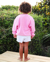 A child standing on a wooden deck outdoors, viewed from behind, wearing a pink long-sleeve sweatshirt and white shorts with curly brown hair.