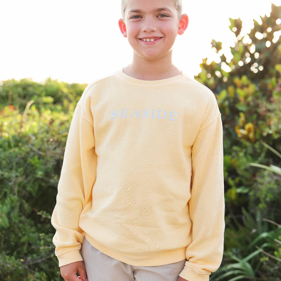 A young boy smiling outdoors wearing a pale yellow long-sleeve crewneck sweatshirt with 'SEASIDE' printed in light blue across the chest, paired with light khaki pants.