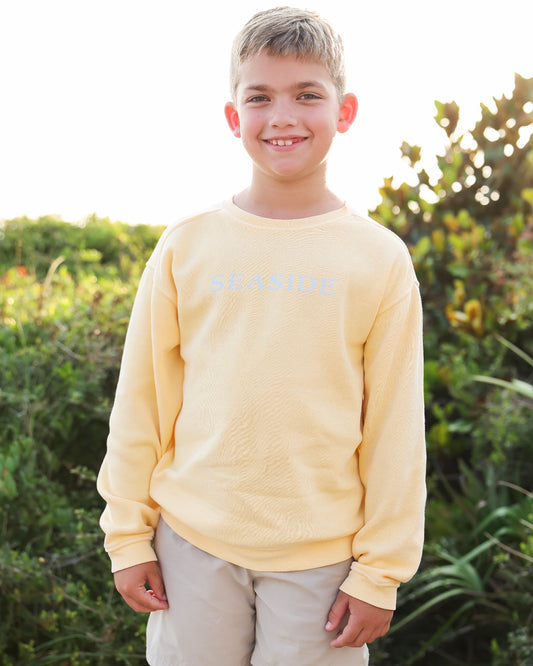 A young boy smiling outdoors wearing a pale yellow long-sleeve crewneck sweatshirt with 'SEASIDE' printed in light blue across the chest, paired with light khaki pants.