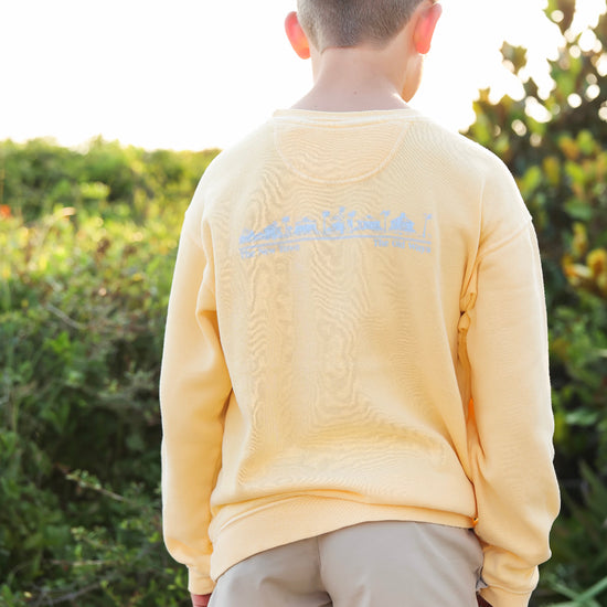 A child viewed from behind wearing a pale yellow sweatshirt with a light blue graphic across the upper back, standing outdoors in front of green foliage.