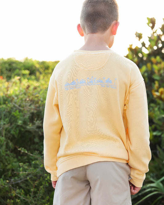 A child viewed from behind wearing a pale yellow sweatshirt with a light blue graphic across the upper back, standing outdoors in front of green foliage.