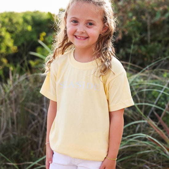 A young girl with blonde hair stands outdoors wearing a pale yellow t-shirt with 'SEASIDE' printed in light blue across the chest, paired with white shorts.