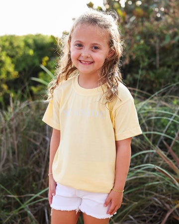 A young girl with blonde hair stands outdoors wearing a pale yellow t-shirt with 'SEASIDE' printed in light blue across the chest, paired with white shorts.
