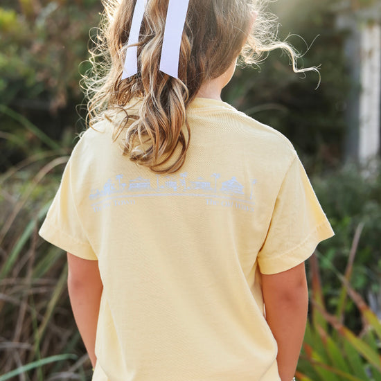 A child viewed from behind wearing a pale yellow t-shirt with a light blue graphic print on the back, set against a blurred outdoor background.