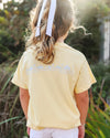 A child viewed from behind wearing a pale yellow t-shirt with a light blue graphic print on the back, set against a blurred outdoor background.