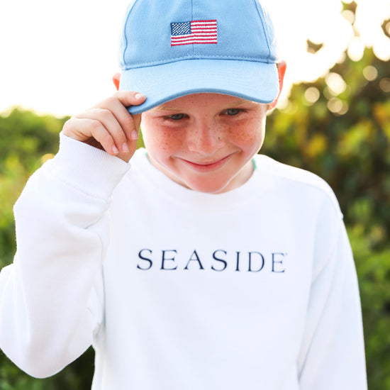 A young boy wearing a light blue baseball cap with an American flag patch and a white long-sleeve sweatshirt with 'SEASIDE' printed on the chest, set against a blurred green outdoor background.