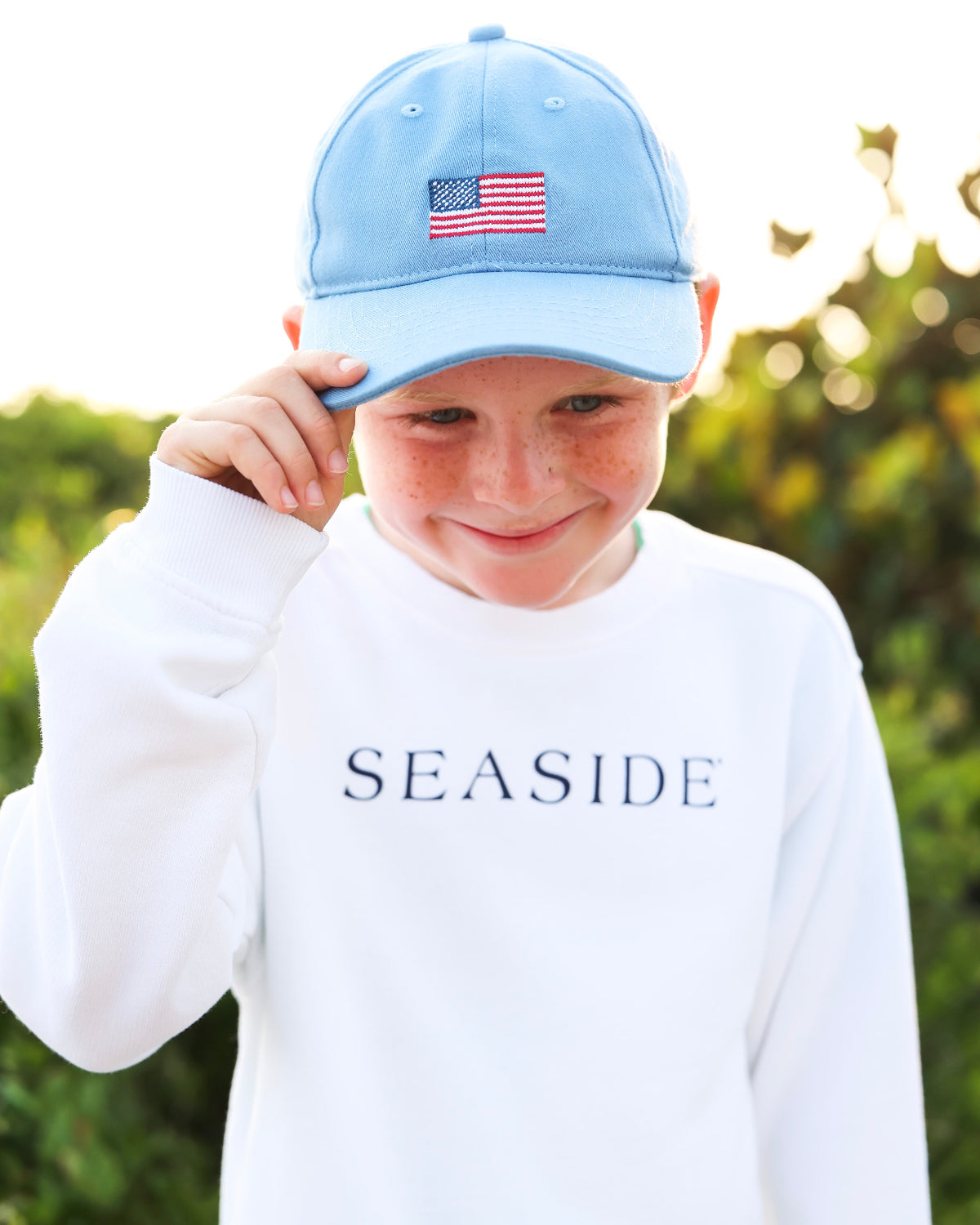 A young boy wearing a light blue baseball cap with an American flag patch and a white long-sleeve sweatshirt with 'SEASIDE' printed on the chest, set against a blurred green outdoor background.