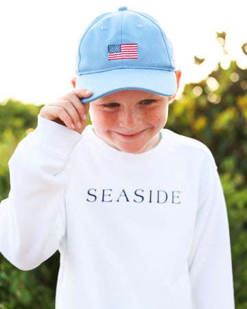 A young boy wearing a light blue baseball cap with an American flag patch and a white long-sleeve sweatshirt with 'SEASIDE' printed on the chest, set against a blurred green outdoor background.