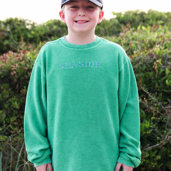 A young boy wearing a bright green corded crewneck ong-sleeve sweatshirt with 'SEASIDE' embroidered on the chest, paired with a navy blue baseball cap that also says 'SEASIDE'. He is standing outdoors in front of a soft-focus background of green beach shrubs.