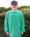 A young boy wearing a bright green corded crewneck ong-sleeve sweatshirt with 'SEASIDE' embroidered on the chest, paired with a navy blue baseball cap that also says 'SEASIDE'. He is standing outdoors in front of a soft-focus background of green beach shrubs.