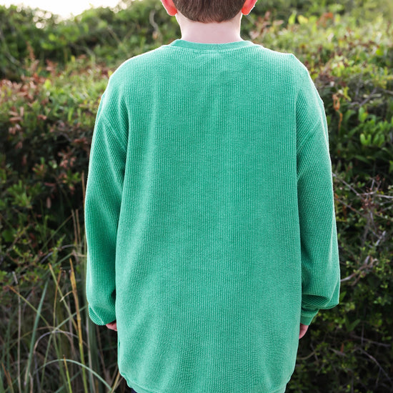 A boy viewed from behind wearing a bright green corded crewneck long-sleeve sweatshirt and a dark baseball cap, standing outdoors in front of green bushes.